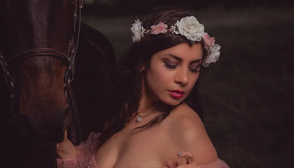 A woman wearing a floral headband poses contemplatively beside a horse in a dimly lit artistic portrait