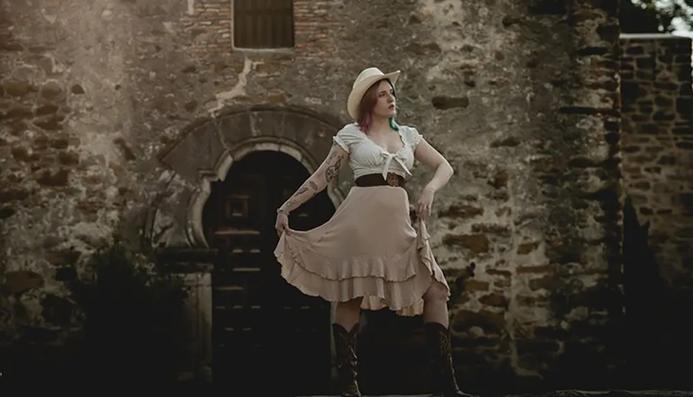 A person in Western-inspired clothing poses confidently in front of an aged stone building exuding a rustic charm