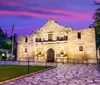 The image shows the historic Alamo Mission in San Antonio Texas at twilight with its facade beautifully illuminated and reflected on a water surface in the foreground