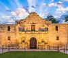 The image shows the historic Alamo Mission in San Antonio Texas at twilight with its facade beautifully illuminated and reflected on a water surface in the foreground