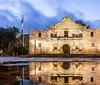 The image shows the historic Alamo Mission in San Antonio Texas at twilight with its facade beautifully illuminated and reflected on a water surface in the foreground