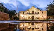 The image shows the historic Alamo Mission in San Antonio, Texas, at twilight, with its facade beautifully illuminated and reflected on a water surface in the foreground.