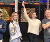 Three friends are enjoying a fun moment in a bar with one of them playfully holding a dart mid-throw while another holds a cocktail all surrounded by various bar signage and a person in the background