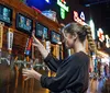 Three friends are enjoying a fun moment in a bar with one of them playfully holding a dart mid-throw while another holds a cocktail all surrounded by various bar signage and a person in the background