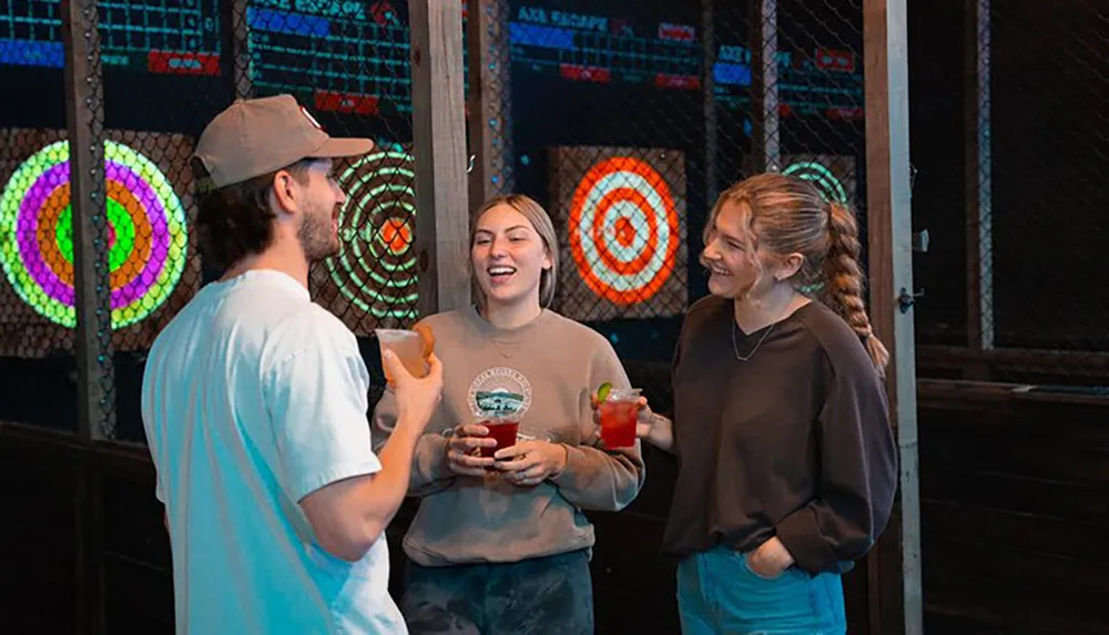 Three people are laughing and enjoying drinks in a social setting with colorful illuminated dartboards in the background