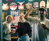 Three friends are enjoying a fun moment in a bar with one of them playfully holding a dart mid-throw while another holds a cocktail all surrounded by various bar signage and a person in the background