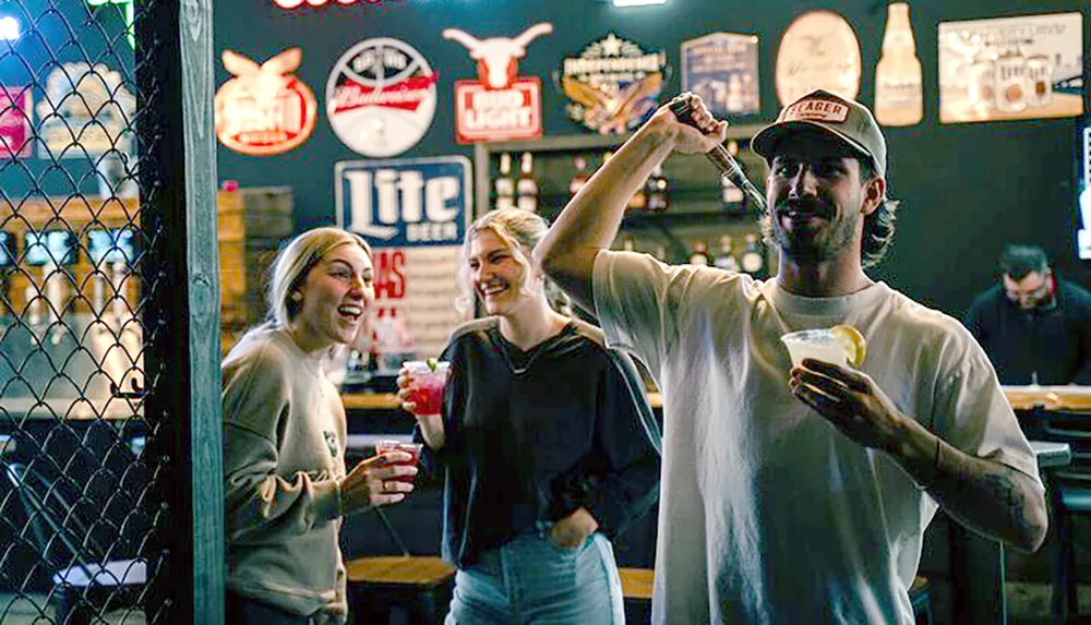 Three friends are enjoying a fun moment in a bar with one of them playfully holding a dart mid-throw while another holds a cocktail all surrounded by various bar signage and a person in the background