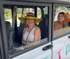 Two smiling individuals are seated inside a small white electric vehicle with an open door the one on the left wearing a colorful wide-brimmed hat