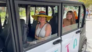 Two smiling individuals are seated inside a small white electric vehicle with an open door, the one on the left wearing a colorful wide-brimmed hat.