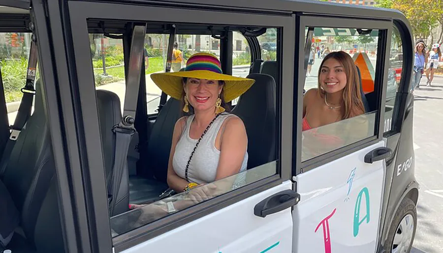 Two smiling individuals are seated inside a small white electric vehicle with an open door, the one on the left wearing a colorful wide-brimmed hat.