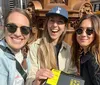 Three smiling women are taking a selfie on a sunny day in an urban setting with one of them holding a book titled Im Glad My Mom Died