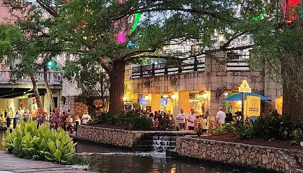 The image captures a lively evening scene with people gathered along a riverwalk with ambient lighting trees and an urban backdrop