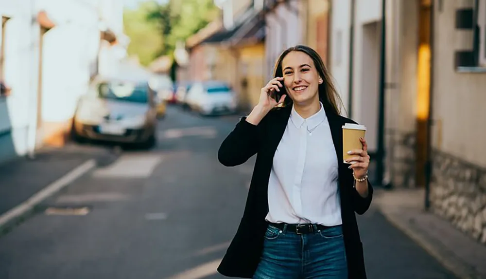 A smiling person is walking down a city street while talking on a mobile phone and holding a coffee cup