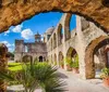 This image shows a view of an old stone mission with arches a dome and a bell tower framed by a weathered stone archway under a clear blue sky