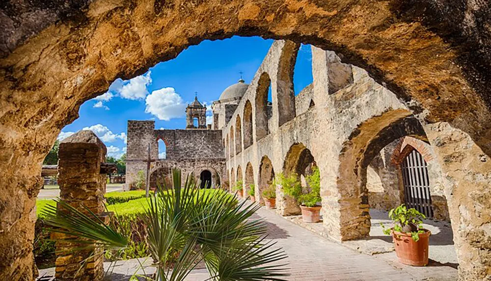 This image shows a view of an old stone mission with arches a dome and a bell tower framed by a weathered stone archway under a clear blue sky