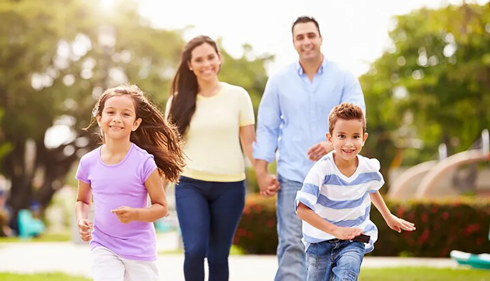 A happy family with two adults and two children are enjoying a sunny day outdoors in the park with the kids running ahead and the parents following behind all smiling