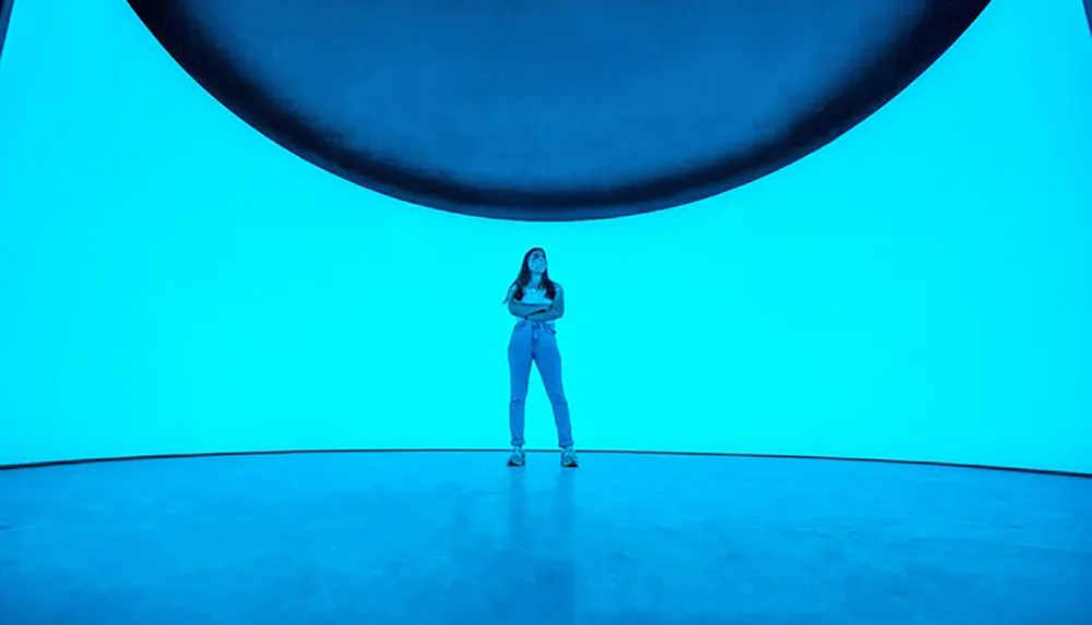 A person stands in the center of a room with blue lighting and a large concave ceiling feature looming above