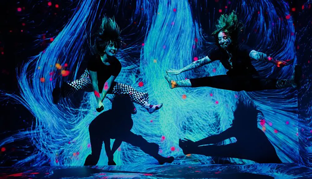 Two dancers are captured in mid-air against a vibrant blue and pink neon-lit background creating a dynamic sense of movement and energy