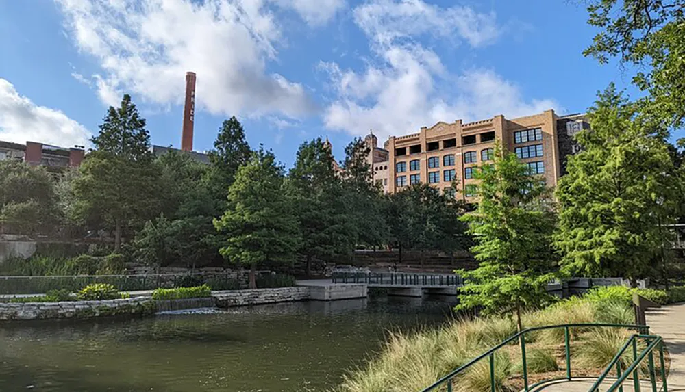 The image captures a serene urban park with a pond green trees a walking bridge and historical industrial buildings in the background under a blue sky with scattered clouds