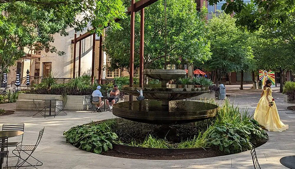 A person in a yellow dress is walking past a tiered fountain in a peaceful urban park setting with seated individuals and lush greenery