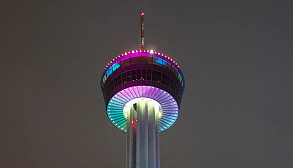 The image shows a lit-up tower at night with a colorful neon-lit circular observation deck