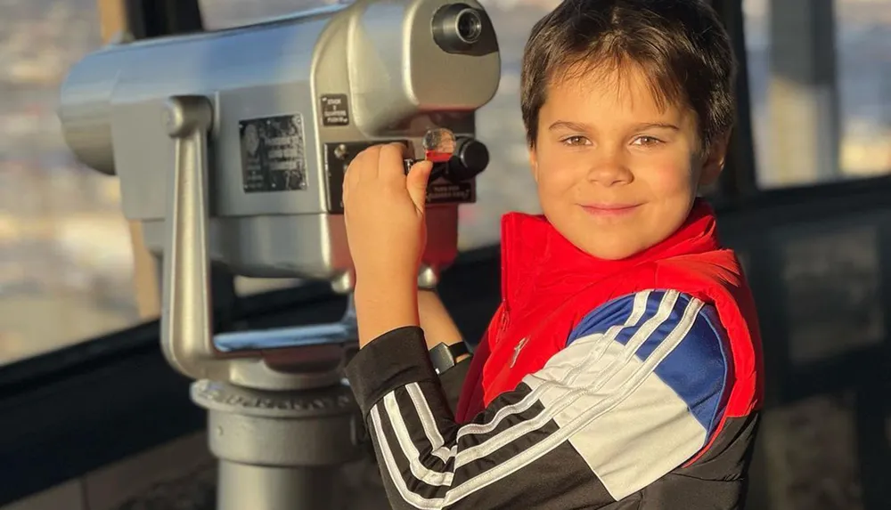 A smiling child stands next to a coin-operated viewing telescope possibly at a scenic overlook or observation deck