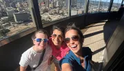 Three people are smiling for a selfie with a cityscape visible from a high vantage point in the background.