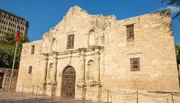 The image shows the historic Alamo mission in San Antonio, Texas, with its distinctive facade under a bright blue sky.