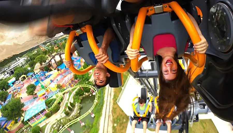 Two people are upside down on a roller coaster, expressing excitement and thrill, with an amusement park visible in the background.