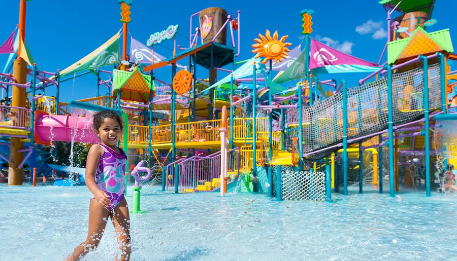 A child is smiling and playing with a water toy in front of a colorful water playground on a sunny day.