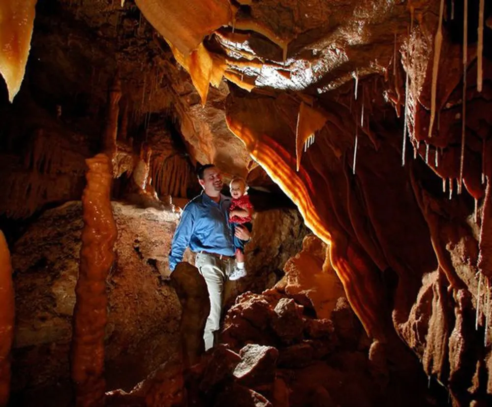 A person is holding a child while exploring an illuminated richly textured cave with stalactites and stalagmites