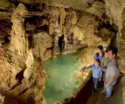 A family is admiring an underground pool in a beautifully lit cave with intricate rock formations surrounding them.