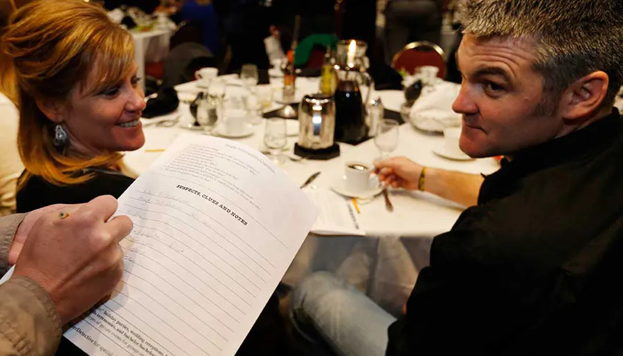 A man and a woman are sharing a moment of engagement over a document at a dining event, with the woman holding a paper and both showing expressions of interest and amusement.