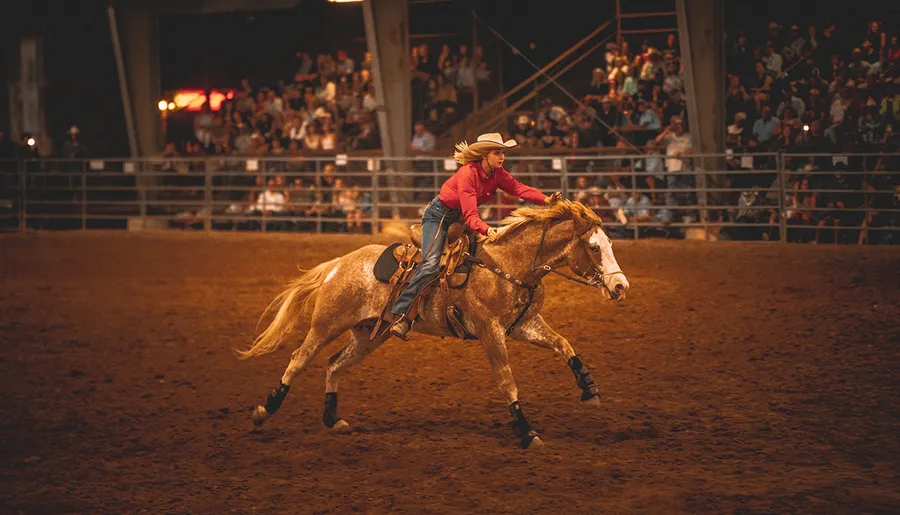 A person in a cowboy hat is riding a horse at a rodeo event, with a crowd watching in the background.