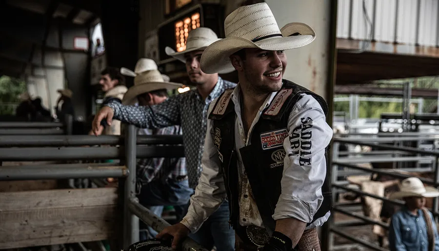 A man in a cowboy hat and rodeo gear smiles while leaning on a fence, with others in similar attire in the background.