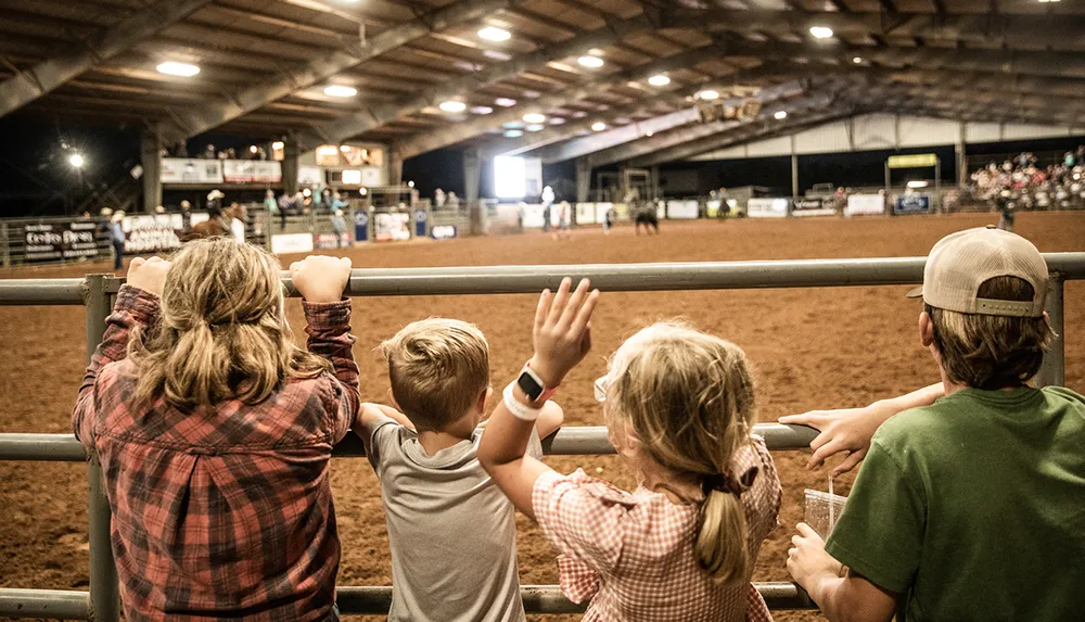 Four children watch a rodeo event from behind a railing inside an arena