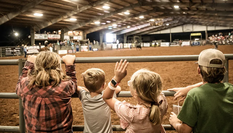 Four children watch a rodeo event from behind a railing inside an arena.