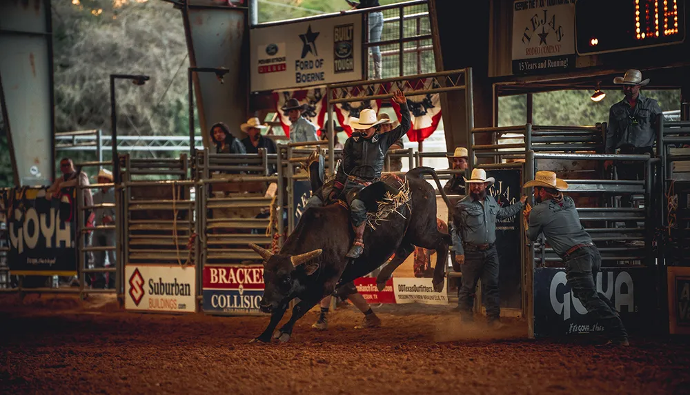 A rodeo rider is mounted on a bucking bull inside an arena surrounded by spectators and rodeo personnel