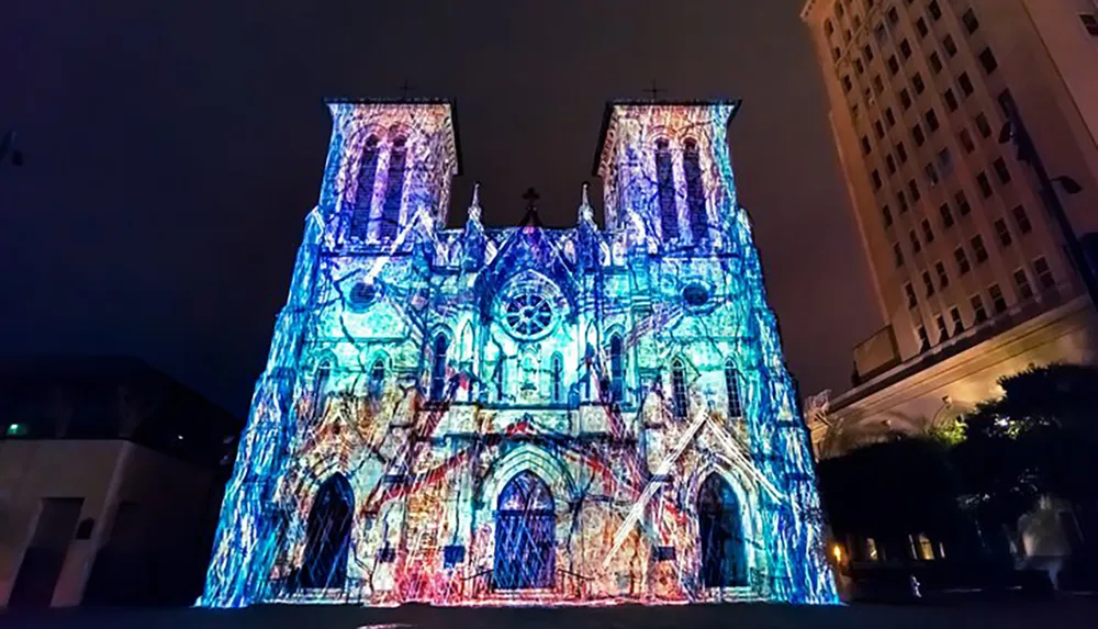 The image shows a cathedral illuminated with a vibrant light projection creating a striking visual display against the night sky