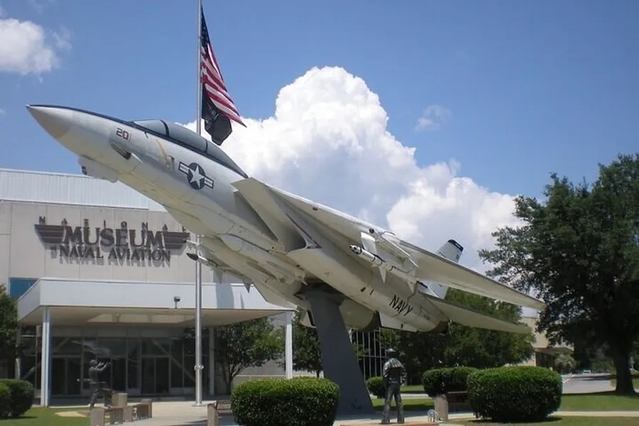 A military fighter jet is on display outside the National Naval Aviation Museum under a clear sky with puffy white clouds.