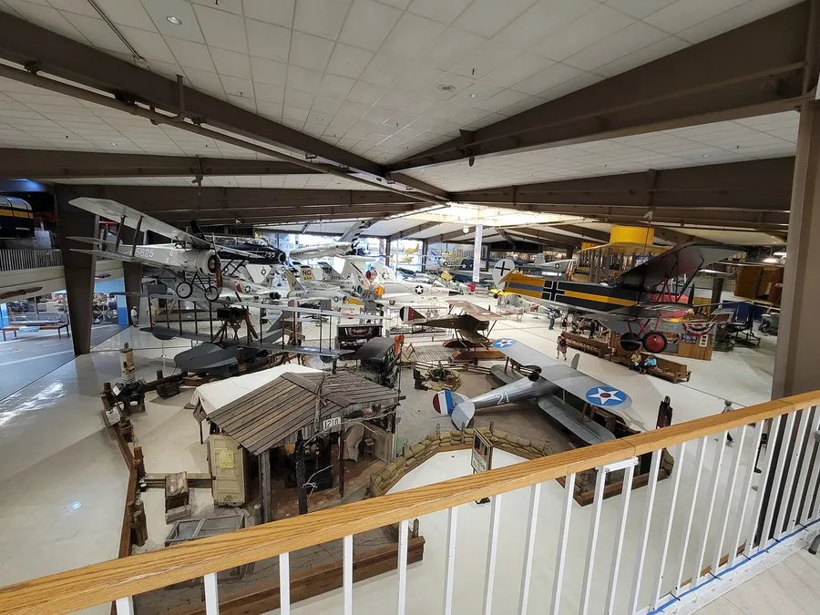 The image shows an aerial view of an expansive indoor museum exhibit featuring a diverse collection of historic airplanes and aviation-related displays.