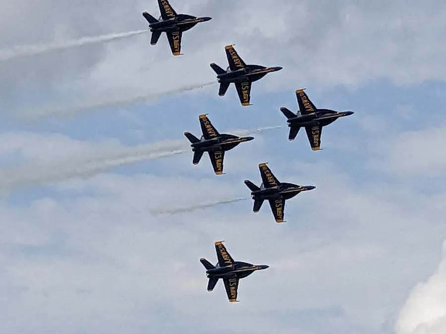 A formation of six blue and yellow jets flying in close proximity with white smoke trails against a cloudy sky.