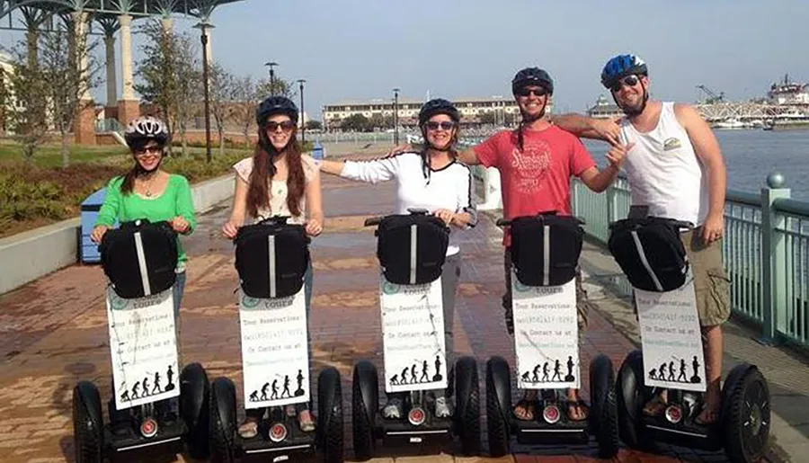 A group of five people are standing on a waterfront boardwalk, smiling and posing with their Segways.