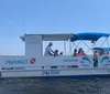 A group of individuals is enjoying a sunny day on a small pontoon boat against the backdrop of coastal buildings and clear blue skies