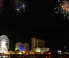 A nighttime cityscape featuring a lit Ferris wheel and brightly colored buildings is shown below a dark sky where multiple fireworks are displayed in separate insets