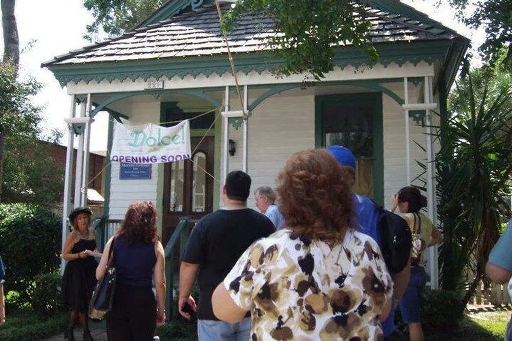 A group of people is standing in front of a quaint house with a sign that reads Hotel OPENING SOON