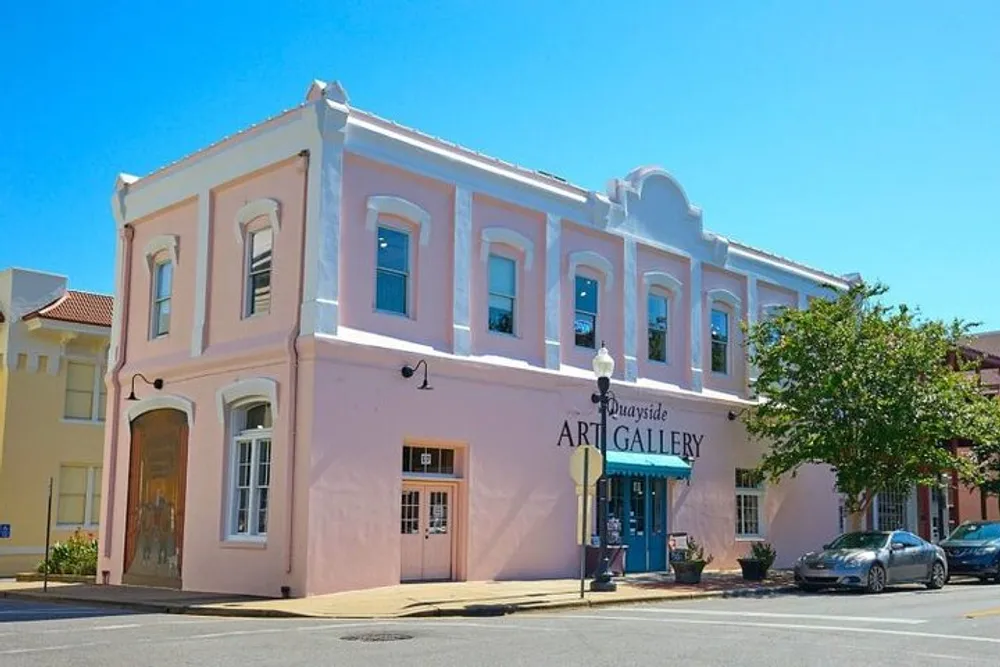 The image shows a pale pink two-story building on a street corner with signage indicating its an art gallery under a clear blue sky