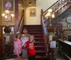 Three people are smiling for a photo at the bottom of a wooden staircase in a vintage-styled interior with walls adorned with framed pictures