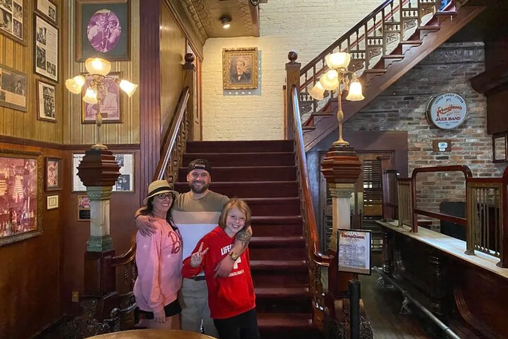 Three people are smiling for a photo at the bottom of a wooden staircase in a vintage-styled interior with walls adorned with framed pictures