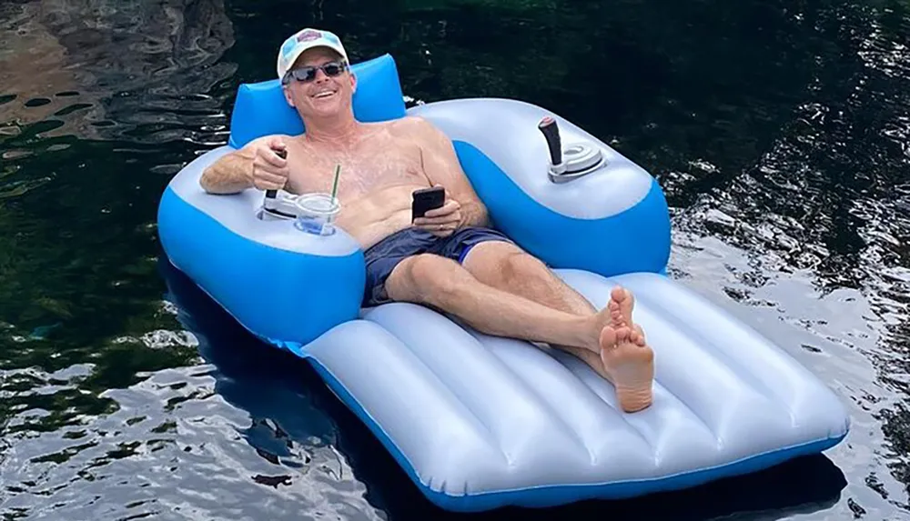 A person is relaxing on a blue and white inflatable raft in the water wearing sunglasses while holding a drink in one hand and a smartphone in the other